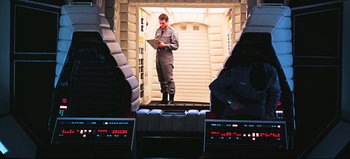 Movie still from “2001: A Space Odyssey” (1968), directed by Stanley Kubrick – A man standing on a stair case holding a laptop; Wide shot, High angle