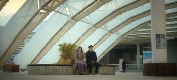 Movie still from “20th Century Girl” (2022), directed by Woo-ri Bang – A man and a woman sitting on a bench in front of a building; Wide shot, High angle