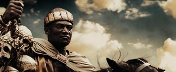 Movie still from “300” (2006), directed by Zack Snyder – A man in a native american costume with a dog in the background; Close Up shot, Low angle