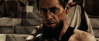 Movie still from “300” (2006), directed by Zack Snyder – An old man with a beard and a mustache is holding something in his mouth; Close Up shot, Low angle