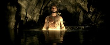 Movie still from “300: Rise of an Empire” (2014), directed by Noam Murro – A man with a bare torso sitting in a pool of water in front of a cave; Medium shot, High angle