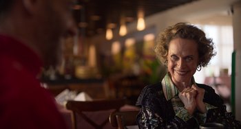 Movie still from “40 Years Young” (2022), directed by Pietro Loprieno – An older woman sitting at a table in a restaurant; Close Up shot, Over the shoulder angle