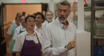 Movie still from “40 Years Young” (2022), directed by Pietro Loprieno – An older man standing in front of a group of people; Medium shot, Over the shoulder angle