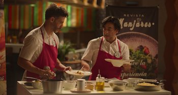 Movie still from “40 Years Young” (2022), directed by Pietro Loprieno – Two men in red aprons preparing food in a kitchen; Medium shot, High angle