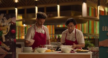 Movie still from “40 Years Young” (2022), directed by Pietro Loprieno – A couple of men standing next to each other in front of bowls of food; Medium shot, Over the shoulder angle