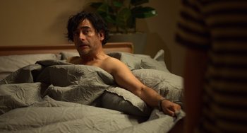 Movie still from “40 Years Young” (2022), directed by Pietro Loprieno – A man laying in a bed under a comforter; Medium shot, High angle