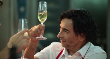 Movie still from “40 Years Young” (2022), directed by Pietro Loprieno – A man holding up a glass of white wine; Close Up shot, Over the shoulder angle