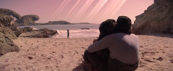 Movie still from “65” (2023), directed by Bryan Woods – Two people sitting on the beach watching a person in the water; Wide shot, High angle
