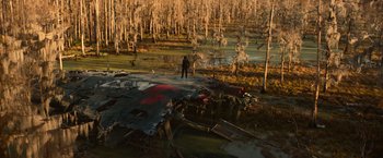 Movie still from “65” (2023), directed by Bryan Woods – A man standing on a dirt road near a body of water; Extreme Wide shot, High angle