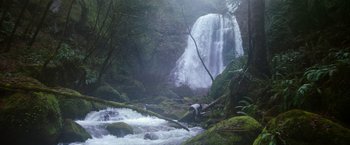 Movie still from “65” (2023), directed by Bryan Woods – A person standing on a tree in front of a waterfall; Extreme Wide shot, Low angle