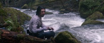 Movie still from “65” (2023), directed by Bryan Woods – A man sitting on a rock near a body of water; Medium shot, Over the shoulder angle