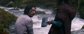 Movie still from “65” (2023), directed by Bryan Woods – A man with long black hair is holding a book; Medium shot, Over the shoulder angle