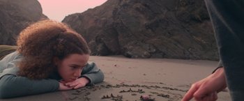 Movie still from “65” (2023), directed by Bryan Woods – A young girl laying in the sand on the beach; Close Up shot, High angle