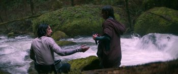 Movie still from “65” (2023), directed by Bryan Woods – Two people are standing near a river and one is holding a bird; Wide shot, Over the shoulder angle