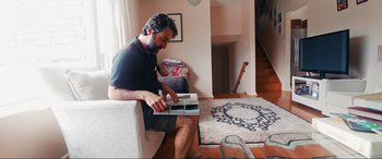 Movie still from “752 Is Not a Number” (2022), directed by Babak Payami – A man sitting on a chair holding a book; Wide shot, Low angle