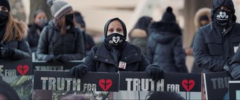 Movie still from “752 Is Not a Number” (2022), directed by Babak Payami – A woman wearing a mask and holding a sign; Medium shot, High angle