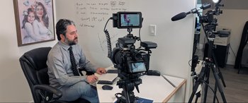 Movie still from “752 Is Not a Number” (2022), directed by Babak Payami – A man sitting in front of a video camera on top of a table; Medium shot, Over the shoulder angle
