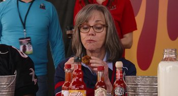 Movie still from “80 for Brady” (2023), directed by Kyle Marvin – A woman eating a hot dog while sitting at a table with many condiments on it; Close Up shot, High angle