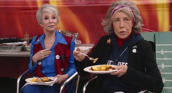 Movie still from “80 for Brady” (2023), directed by Kyle Marvin – Two older women sitting next to each other holding plates of food; Medium shot, High angle