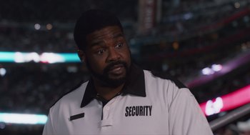 Movie still from “80 for Brady” (2023), directed by Kyle Marvin – A man in a white security uniform looking at the camera; Medium shot, High angle