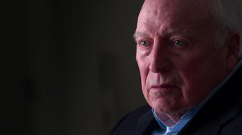 Movie still from “9/11: Inside the President's War Room” (2021), directed by Adam Wishart – An older man in a black suit and blue shirt; Close Up shot, Low angle