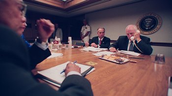 Movie still from “9/11: Inside the President's War Room” (2021), directed by Adam Wishart – A group of people sitting at a table with food; Medium shot, Over the shoulder angle