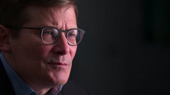 Movie still from “9/11: Inside the President's War Room” (2021), directed by Adam Wishart – A person wearing glasses and a suit; Close Up shot, Low angle