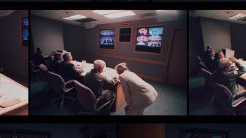 Movie still from “9/11: Inside the President's War Room” (2021), directed by Adam Wishart – A group of people sitting at a table in front of a tv; Wide shot, High angle