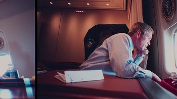 Movie still from “9/11: Inside the President's War Room” (2021), directed by Adam Wishart – A man sleeping on top of a desk next to a suitcase; Medium shot, High angle
