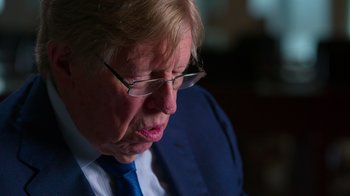 Movie still from “9/11: Inside the President's War Room” (2021), directed by Adam Wishart – An older man wearing glasses and a blue tie; Close Up shot, Over the shoulder angle