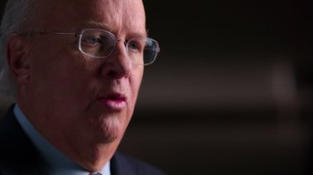 Movie still from “9/11: Inside the President's War Room” (2021), directed by Adam Wishart – An older man wearing glasses and a suit; Close Up shot, Over the shoulder angle