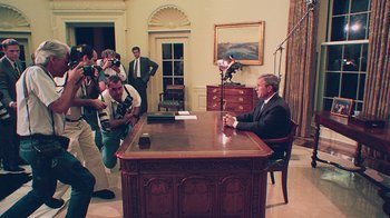 Movie still from “9/11: Inside the President's War Room” (2021), directed by Adam Wishart – A man sitting at a desk in front of a group of people; Wide shot, High angle
