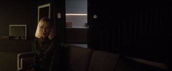 Movie still from “A Banquet” (2021), directed by Ruth Paxton – A dark room with a person sitting on a couch; Medium shot, Over the shoulder angle