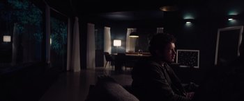 Movie still from “A Banquet” (2021), directed by Ruth Paxton – A man sitting on a couch in a dark room; Medium shot, Over the shoulder angle
