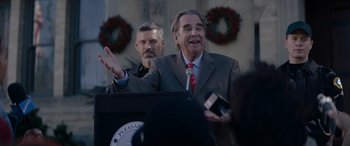 Movie still from “A Christmas Mystery” (2022), directed by Alex Ranarivelo – A man in a gray suit is speaking at a podium; Medium shot, Over the shoulder angle