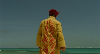 Movie still from “The Fall” (2006), directed by Tarsem Singh – A man in a red hat and a yellow coat stands in front of the ocean; Medium shot, Over the shoulder angle