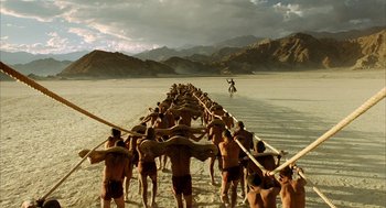 Movie still from “The Fall” (2006), directed by Tarsem Singh – A group of men in swim trunks holding wooden oars in the sand; Extreme Wide shot, High angle