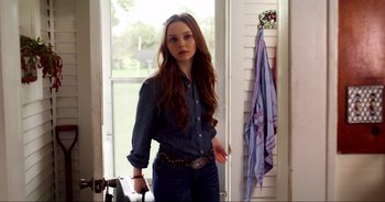 Movie still from “A Cowgirl's Song” (2022), directed by Timothy Armstrong – A woman in a blue shirt is standing by a window; Medium shot, Over the shoulder angle