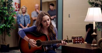 Movie still from “A Cowgirl's Song” (2022), directed by Timothy Armstrong – A woman playing an acoustic guitar in front of a group of people; Medium shot, Over the shoulder angle