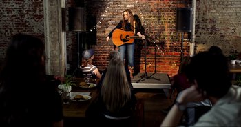 Movie still from “A Cowgirl's Song” (2022), directed by Timothy Armstrong – A woman standing on a stage holding a guitar; Wide shot, Over the shoulder angle