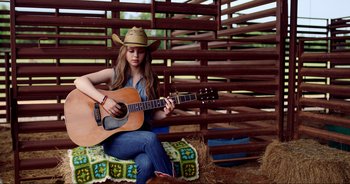 Movie still from “A Cowgirl's Song” (2022), directed by Timothy Armstrong – A woman in a cowboy hat playing an acoustic guitar; Wide shot, Low angle