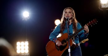 Movie still from “A Cowgirl's Song” (2022), directed by Timothy Armstrong – A woman holding a guitar and singing into a microphone; Medium shot, Over the shoulder angle