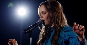 Movie still from “A Cowgirl's Song” (2022), directed by Timothy Armstrong – A girl with long brown hair singing into a microphone; Close Up shot, Over the shoulder angle