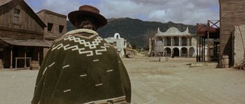 Movie still from “A Fistful of Dollars” (1964), directed by Sergio Leone – A man wearing a blanket and a hat in the middle of an empty street; Wide shot, Over the shoulder angle