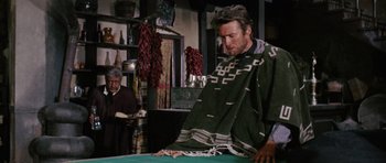 Movie still from “A Fistful of Dollars” (1964), directed by Sergio Leone – A man wearing a green and white blanket standing next to a table; Medium shot, Low angle