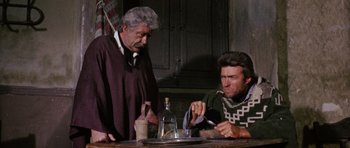 Movie still from “A Fistful of Dollars” (1964), directed by Sergio Leone – Two men sitting at a wooden table with a bottle of wine; Medium shot, Over the shoulder angle