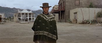 Movie still from “A Fistful of Dollars” (1964), directed by Sergio Leone – A man wearing a poncho and a hat standing in the middle of an empty street; Wide shot, Low angle