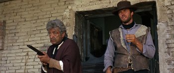 Movie still from “A Fistful of Dollars” (1964), directed by Sergio Leone – Two men standing next to each other in front of a brick building; Medium shot, Low angle