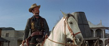 Movie still from “A Fistful of Dollars” (1964), directed by Sergio Leone – A man sitting on top of a white horse next to a wooden fence; Medium shot, Low angle