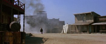 Movie still from “A Fistful of Dollars” (1964), directed by Sergio Leone – A person walking in front of a building with smoke coming out of it; Extreme Wide shot, Low angle